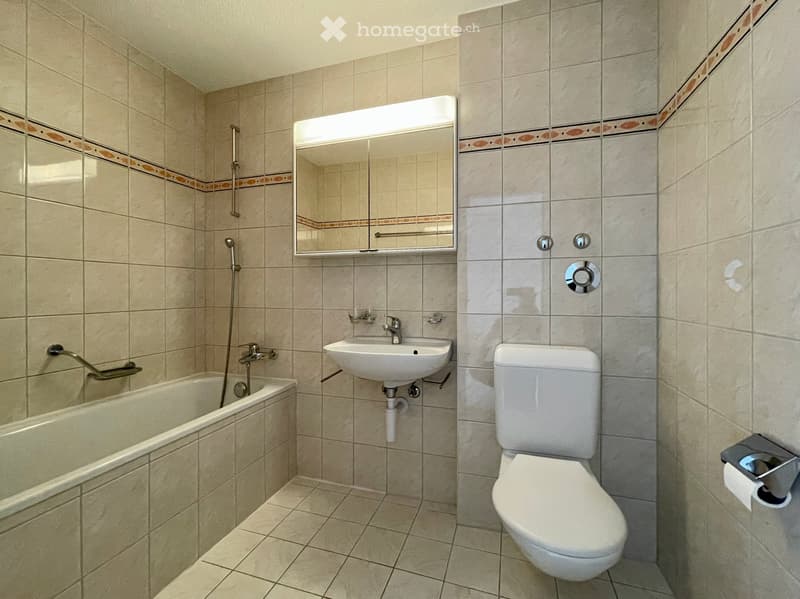 Bathroom with beige tiled walls and floor, featuring a bathtub, sink, toilet, and a mirror above the sink.