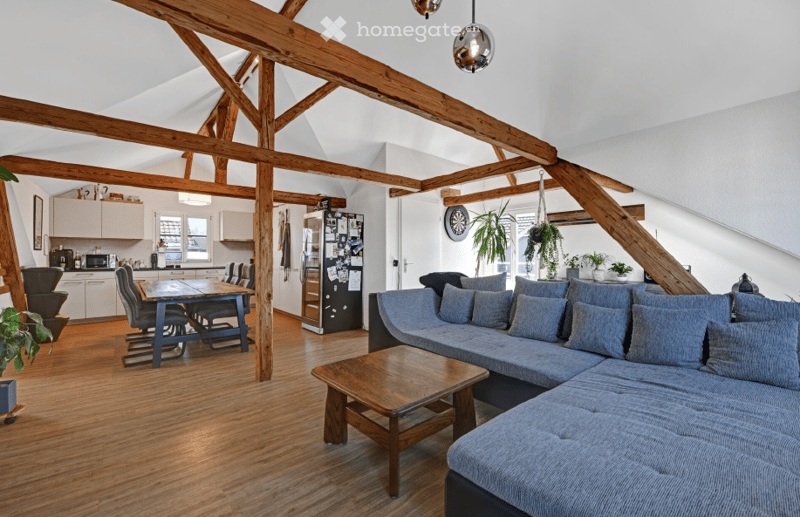 Open-plan living and dining area with exposed wooden beams, grey sectional sofa, wooden coffee table, dining table with chairs, kitchen area with cabinets and appliances.