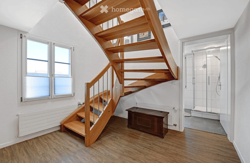 A modern hallway with wooden flooring, a wooden staircase with a metal railing, a small wooden bench, and a glass-enclosed shower area visible in the background.