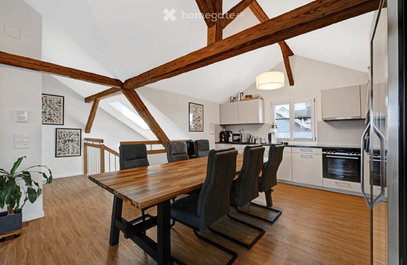 Open-plan dining and kitchen area with exposed wooden beams, a large wooden dining table surrounded by six chairs, modern kitchen cabinets, and a coffee machine on the counter.