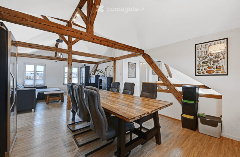 Open-plan living and dining area with exposed wooden beams, hardwood flooring, a large wooden dining table with six chairs, a sofa, coffee table, and a refrigerator. The space is well-lit with large windows and features decorative artwork on the walls.
