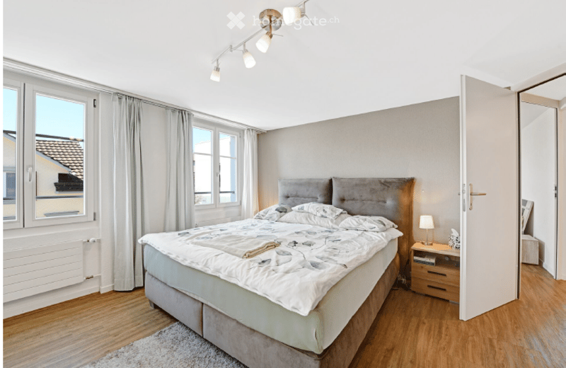 Modern bedroom with a large bed, nightstand, lamp, and curtains. The room features wooden flooring and large windows with a view of the roof.