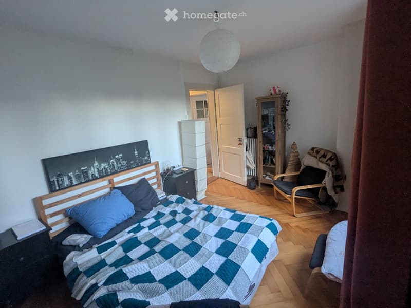 Cozy bedroom with a wooden bed, blue and white checkered quilt, nightstand, chair, cabinet, and a large cityscape picture on the wall.