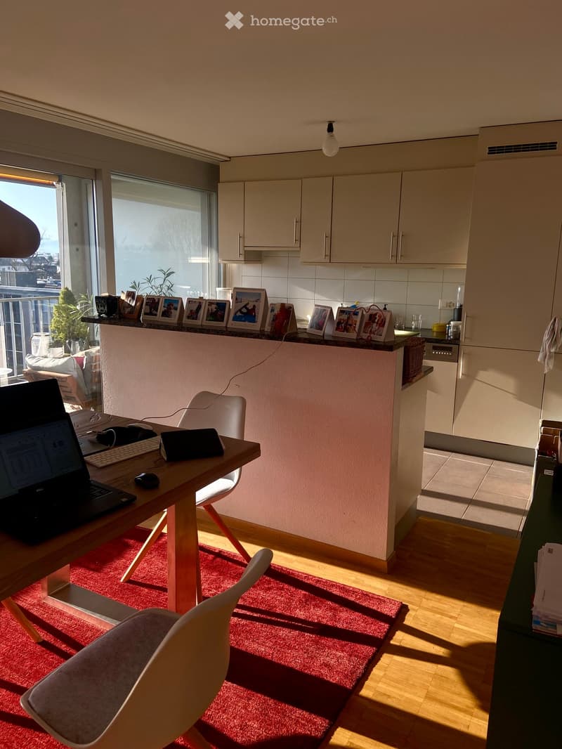 Open-plan living and kitchen area with a wooden table, white chairs, laptop, and framed photos on the counter. The kitchen features white cabinets and a dishwasher, with a balcony visible through large windows.