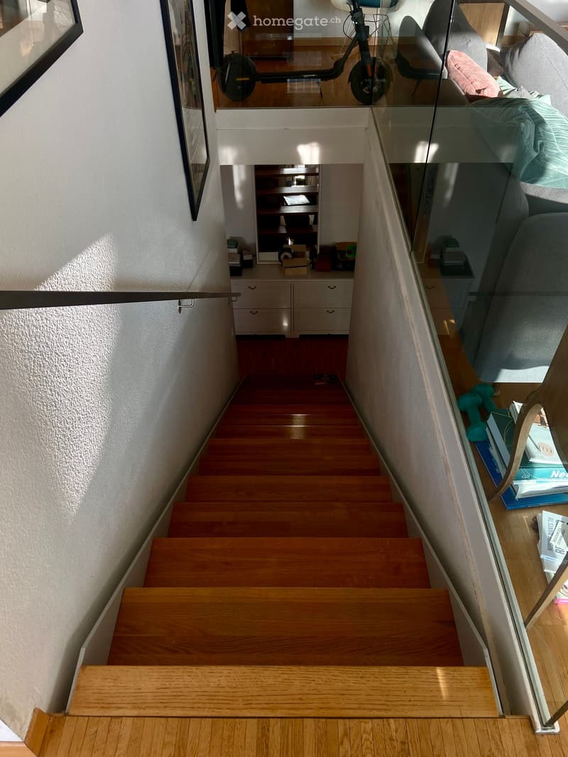 Wooden staircase with a glass railing, leading to a room with a sofa and exercise bike visible in the background.