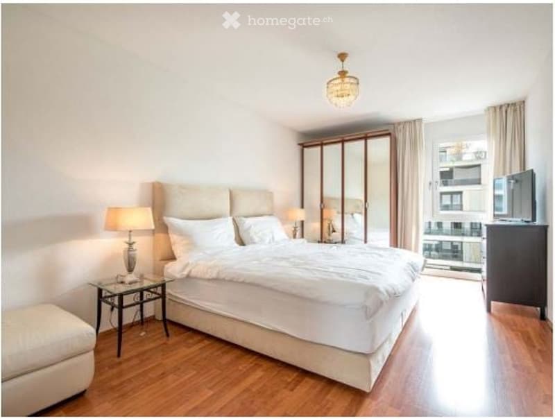 Modern bedroom with a large bed, nightstands, lamps, a chair, a mirror, and a television. The room features wooden flooring and large windows with a view.