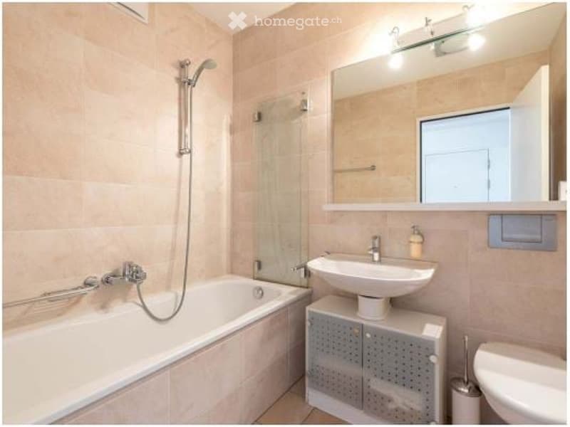 Modern bathroom with a bathtub, sink, mirror, and showerhead. The walls are tiled in a light beige color, and the floor is also tiled. There is a small cabinet under the sink.