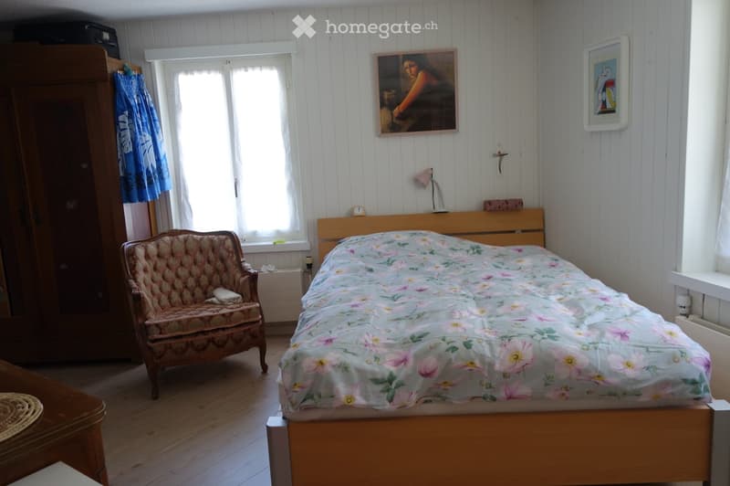 Cozy bedroom with wooden floor, a bed with floral bedding, a wooden chair, a wardrobe, a small table, and a lamp on the wall.