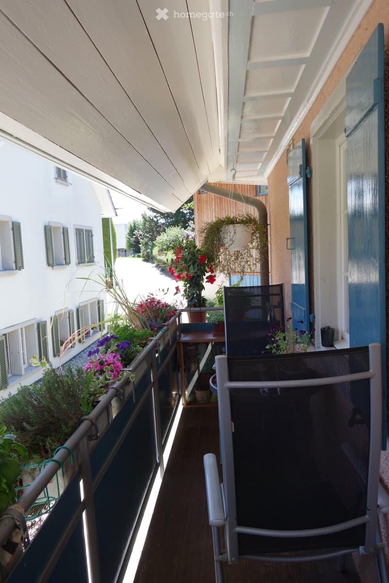 Balcony with wooden floor, metal railing, potted plants, and a small table with a chair.