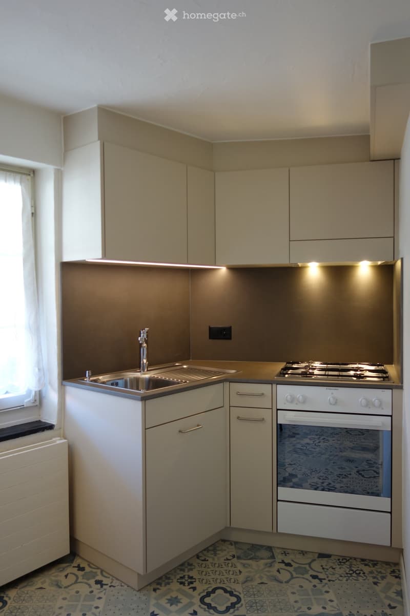 Compact kitchen with light-colored cabinets, a sink, stove, and oven. Features a patterned tile floor and built-in lighting above the counter.