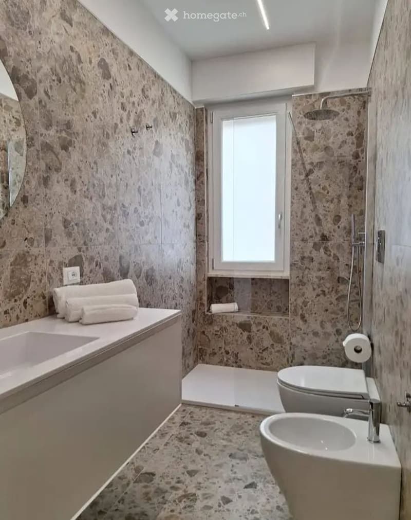 Modern bathroom with stone tile walls and floor, double sink vanity, shower stall, toilet, bidet, and neatly folded towels.