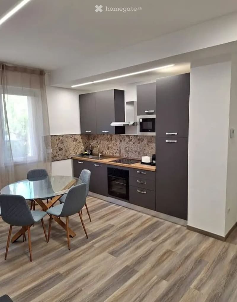 Modern kitchen and dining area with grey cabinets, wooden countertops, a glass dining table with four chairs, and built-in appliances including a microwave, oven, and stove.