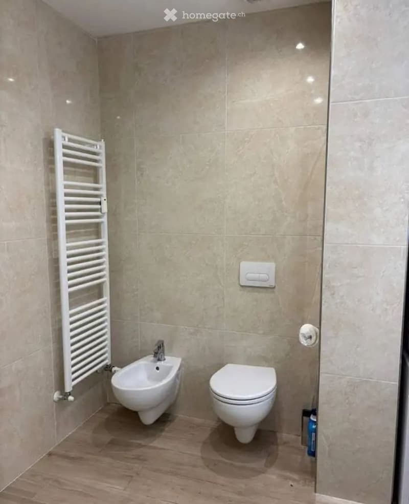 Modern bathroom with beige tiled walls, wooden floor, toilet, bidet, and a white radiator on the left side.