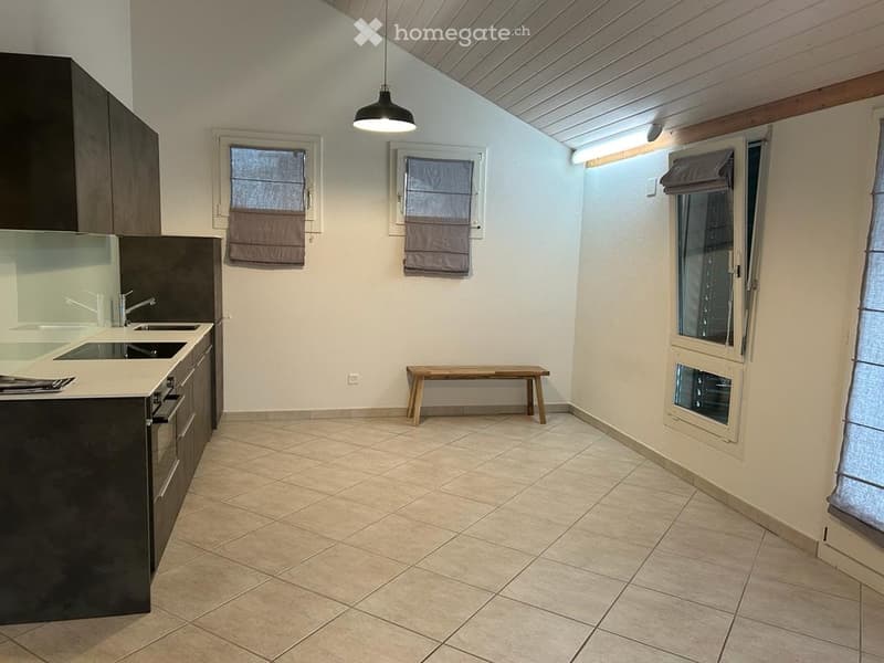 Modern kitchen with dark cabinets, white countertops, a sink, and a small wooden bench. The space features tiled flooring and large windows with blinds.