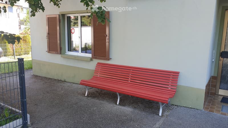 A red wooden bench is placed on a gravel surface next to a house with white walls and wooden shutters. The area is bordered by a metal fence and greenery.