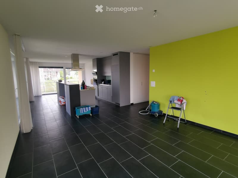Open-plan living and kitchen area with dark tiled flooring, grey cabinets, and a kitchen island. A yellow accent wall is visible, along with a vacuum cleaner, baby chair, and a small cart.