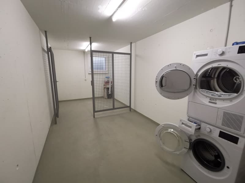 A small, clean room with a washing machine and dryer, white walls, and a metal gate leading to another area.
