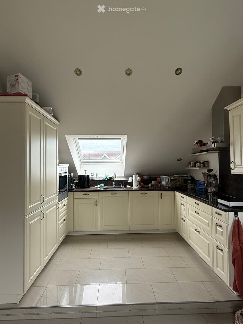 Bright kitchen with cream-colored cabinets, white tiled floor, and a black countertop. Features include a sink with a window above, an oven, microwave, and various small appliances.