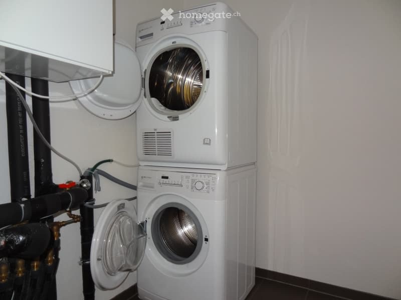 A compact bathroom with a stacked washing machine and dryer unit, white walls, and visible plumbing connections.