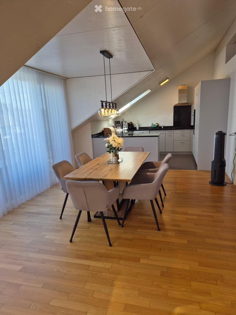 Modern dining area with a wooden dining table, six chairs, a vase with white flowers, and a kitchen area with cabinets and appliances.