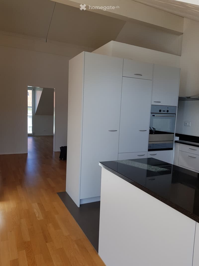 Modern kitchen with white cabinets, black countertop, and wooden flooring. An open area leads to another room with large windows.