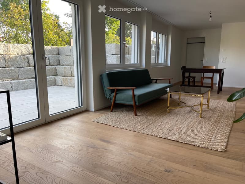 Modern living room with wooden flooring, large windows, a green sofa, a glass coffee table, and a dining area with a table and chairs.