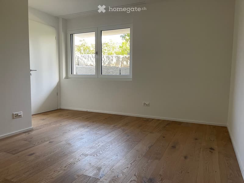 Empty room with wooden flooring, white walls, and a large window with a view of a stone wall and greenery outside.