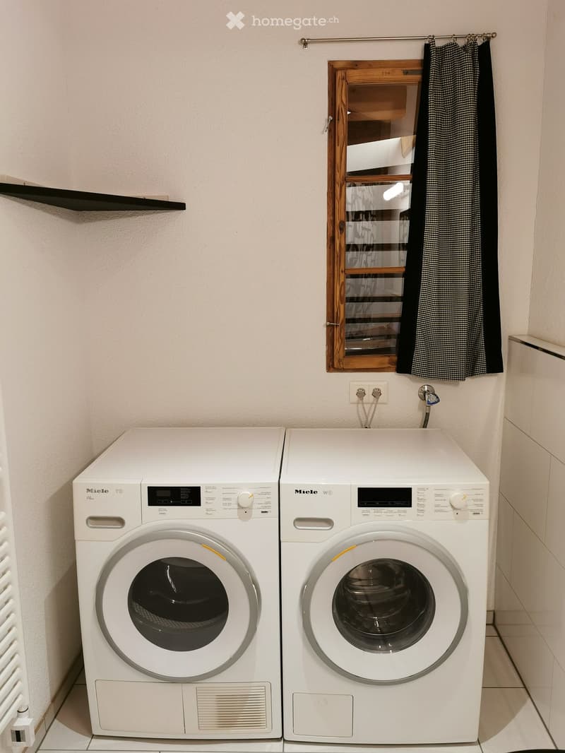 A modern bathroom with a washing machine and dryer side by side, a wooden-framed mirror with a black checkered towel, and a black shelf on the left wall.
