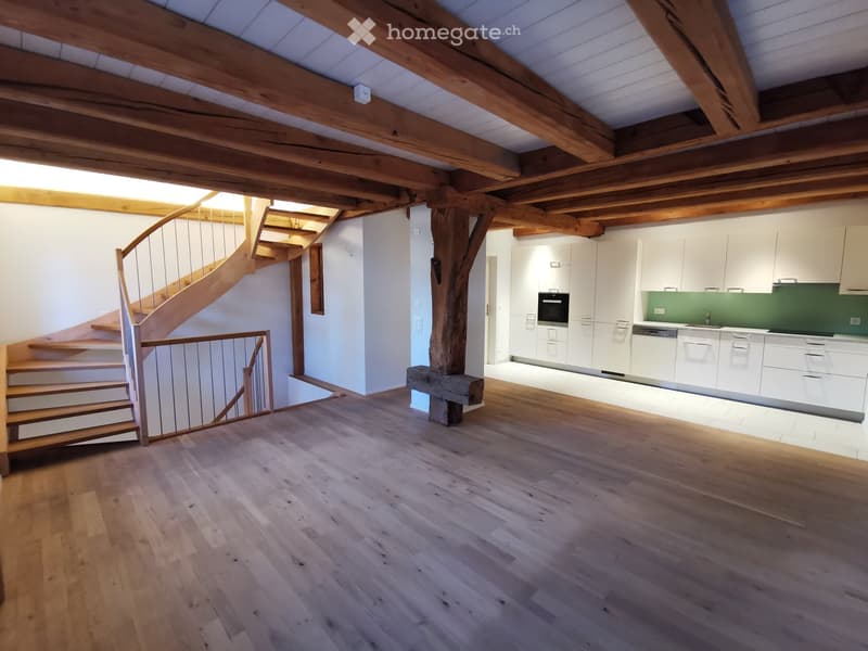 Open-plan space with wooden floors, exposed wooden beams, a staircase leading to an upper level, and a modern kitchen area with white cabinets and stainless steel appliances.