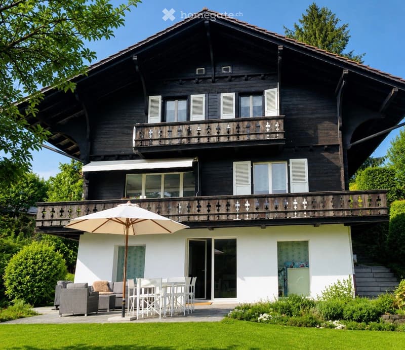 Two-story wooden house with balconies, grey sofa, white dining set, umbrella, and garden.