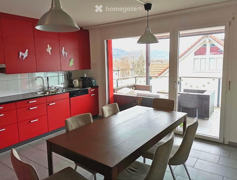 Modern kitchen with red cabinets, a wooden dining table, and chairs, leading to a balcony with a scenic view.