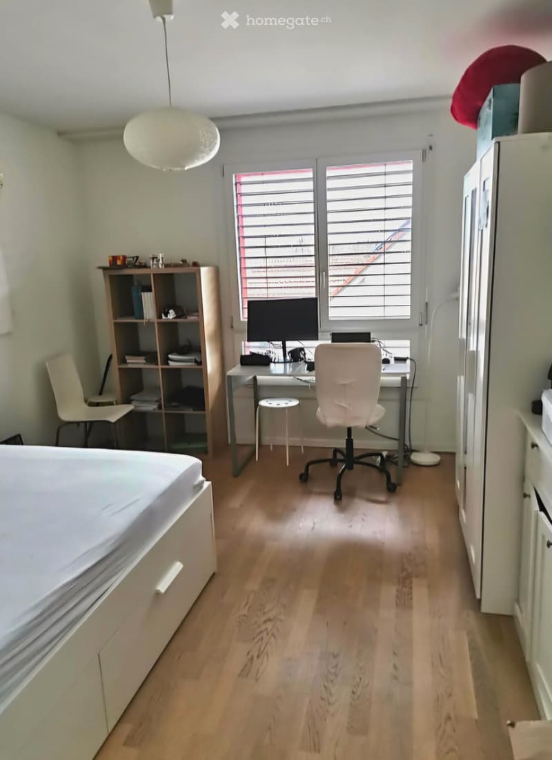 A furnished room with a bed, desk, chair, laptop, screen, shelf, and lamp. The room has wooden flooring and large windows with blinds.