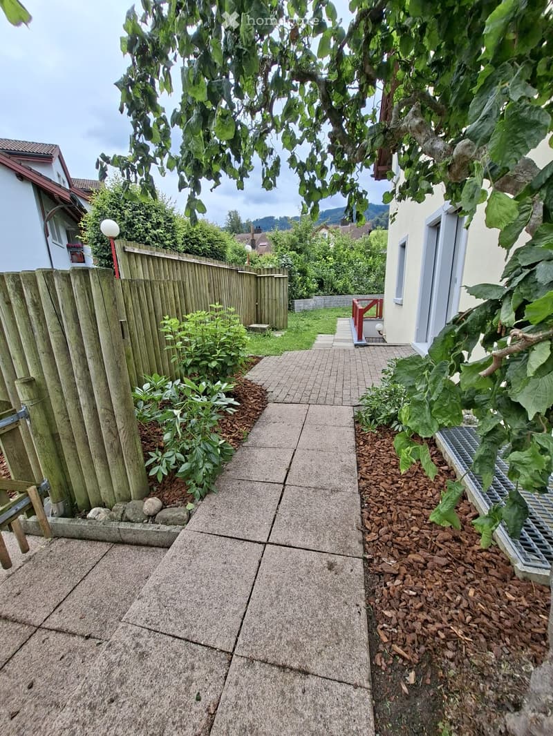 A narrow concrete pathway bordered by a wooden fence on one side and a house on the other, with small plants and mulch beds along the edges.