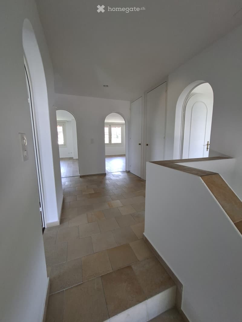 Empty hallway with tiled floor, white walls, arched doorways, and a wooden railing on the right side.