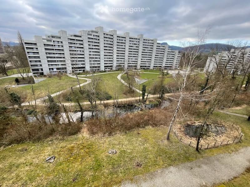 A large residential complex with multiple apartment buildings surrounded by green spaces, pathways, and a small water feature.