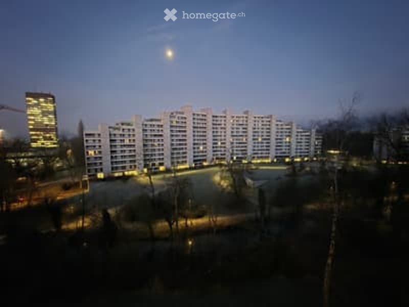 A large residential building with multiple floors and numerous windows, illuminated at night. The surrounding area includes a parking lot and some trees.