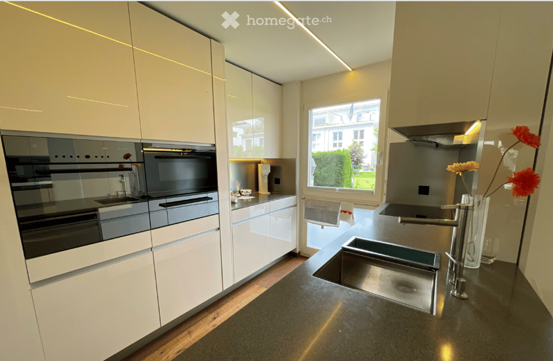 Modern kitchen with white cabinets, stainless steel sink, built-in oven, and a large window offering a view of the garden.