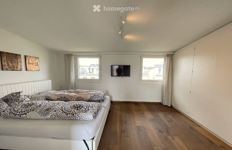 Modern bedroom with a white bed, patterned pillows and blanket, mounted TV, large windows with curtains, and artwork on the walls.