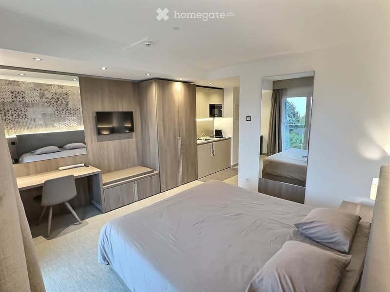 Modern, well-furnished room with a large bed, desk, chair, television, wardrobe, and a small kitchenette area with a sink and microwave.