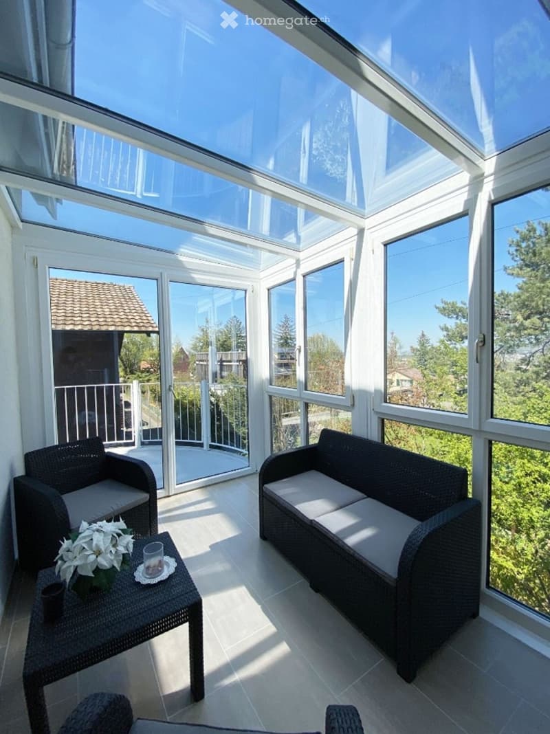 Furnished Room. Bright, airy room with large windows and a skylight, featuring a wicker sofa, wicker chair, small table with a flower arrangement, and a view of the garden and neighboring houses.