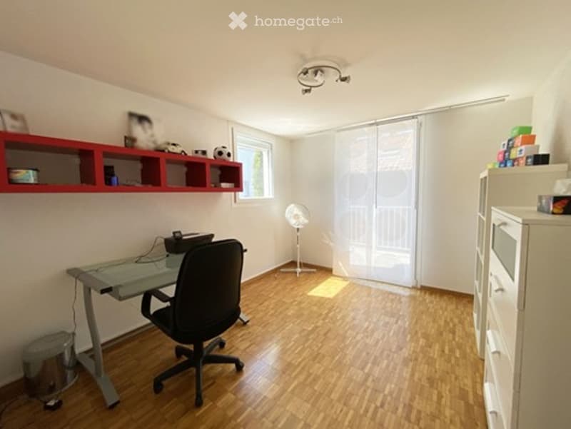 Furnished Room. A modern, well-lit room with wooden flooring, a large window with blinds, a desk with a chair, a red shelf unit, and a standing fan. The room appears to be a home office or study area.