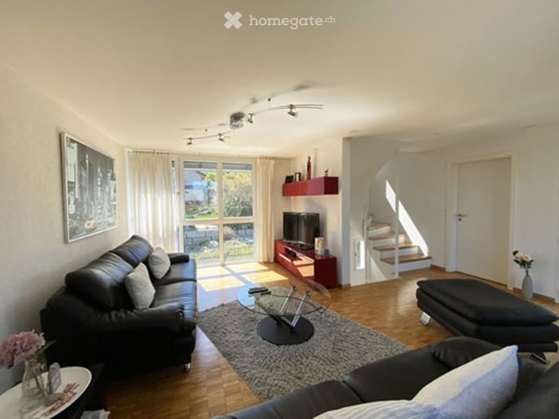 Living Room. Modern living room with hardwood flooring, large windows, black leather sofa, ottoman, glass coffee table, TV stand, and decorative items.