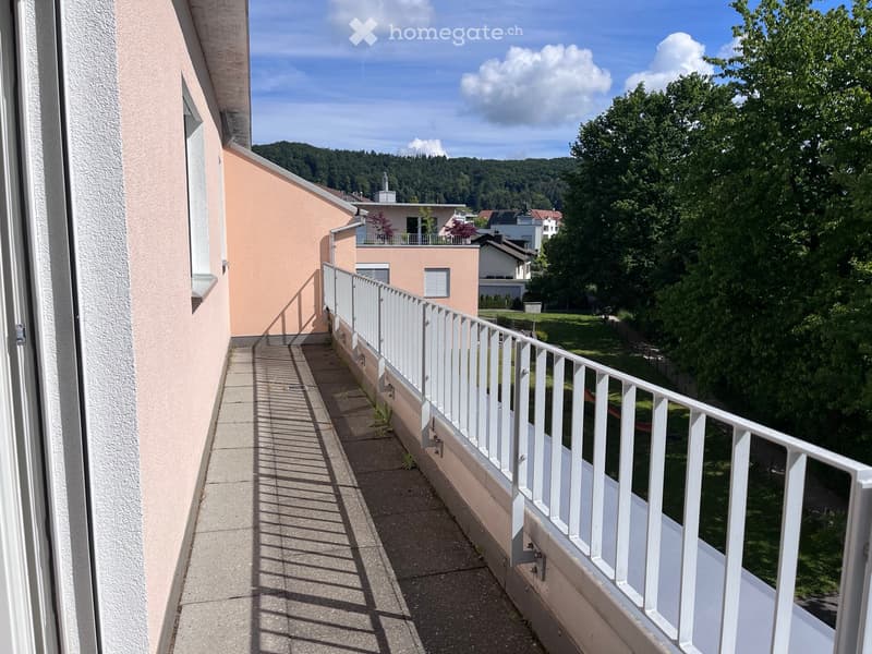 Balcony with white railing, concrete floor, view of greenery and neighboring buildings.