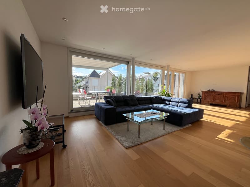 Modern living room with wooden flooring, large glass doors leading to a balcony, a dark blue sectional sofa, a glass coffee table, a small wooden stool with a flower vase, and a flat-screen TV on the wall.