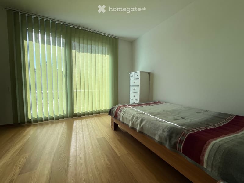 A minimalist bedroom with a wooden bed, a chest of drawers, and vertical blinds covering large windows, allowing natural light to filter in.