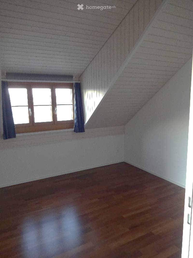 An empty room with white walls, wooden flooring, and a window with blue trim. The ceiling has a slanted design with visible beams.