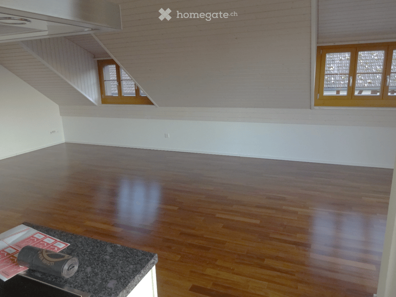 Empty room with wooden flooring, white walls, and large windows with wooden frames. A small table with a rolled-up item and some papers is visible in the foreground.