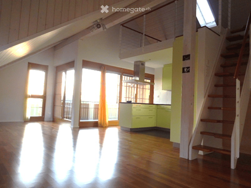 Bright, open-plan space with wooden flooring, large windows, and modern green kitchen units visible in the background. No furniture present.