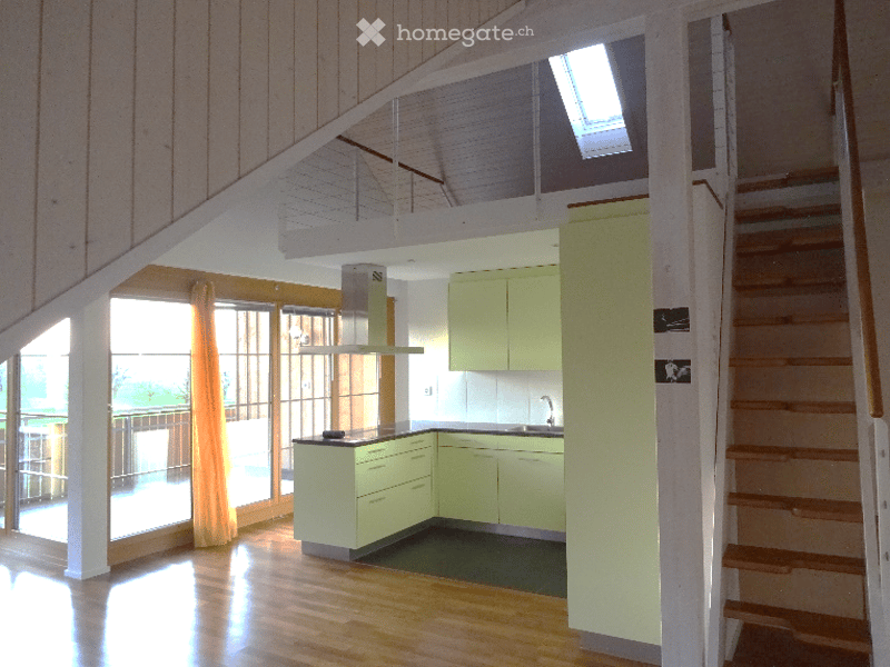 Bright, modern kitchen with light green cabinets, wooden flooring, and a staircase leading to an upper level. Features include a sink, faucet, and large windows with blinds.