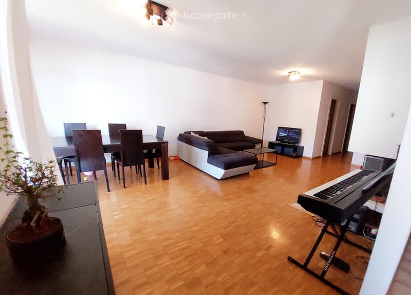 Open-plan living and dining area with wooden flooring, white walls, and large windows. Features a grey sofa with an ottoman, a coffee table, a dining table with chairs, a laptop, a piano, and a TV screen.